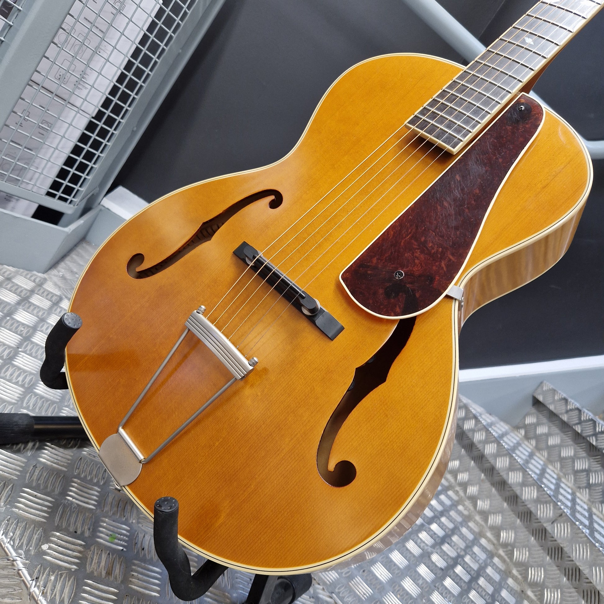 Pre-Owned Epiphone Masterbilt Zenith Classic Electro-Acoustic - Vintage Natural A close-up of a mandolin-style acoustic guitar with a natural wood body, dark f-holes, a dark reddish-brown pickguard, and silver metal bridge hardware, resting on a stand.