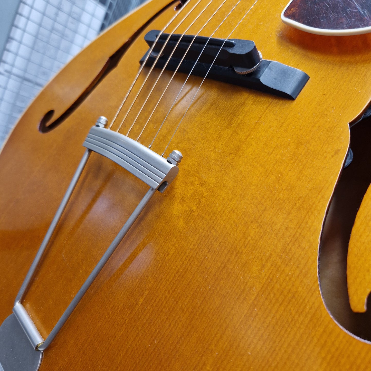 Pre-Owned Epiphone Masterbilt Zenith Classic Electro-Acoustic - Vintage Natural Close-up of an orange-gold f-hole acoustic guitar body with a silver tailpiece and black bridge, photographed outdoors against a blurred building background.