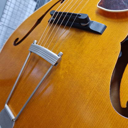 Pre-Owned Epiphone Masterbilt Zenith Classic Electro-Acoustic - Vintage Natural Close-up of an orange-gold f-hole acoustic guitar body with a silver tailpiece and black bridge, photographed outdoors against a blurred building background.