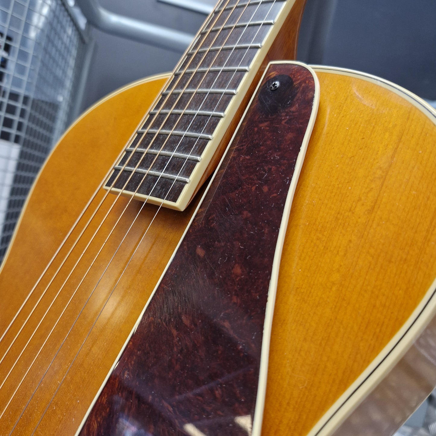 Pre-Owned Epiphone Masterbilt Zenith Classic Electro-Acoustic - Vintage Natural Close-up of an electric guitar body showing the sunburst maple top, dark reddish-brown pickguard, and part of the fretboard with strings.