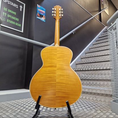 Pre-Owned Epiphone Masterbilt Zenith Classic Electro-Acoustic - Vintage Natural A natural wood acoustic guitar shown from the back, resting on a black stand in an indoor setting with a staircase and dark gray walls in the background.