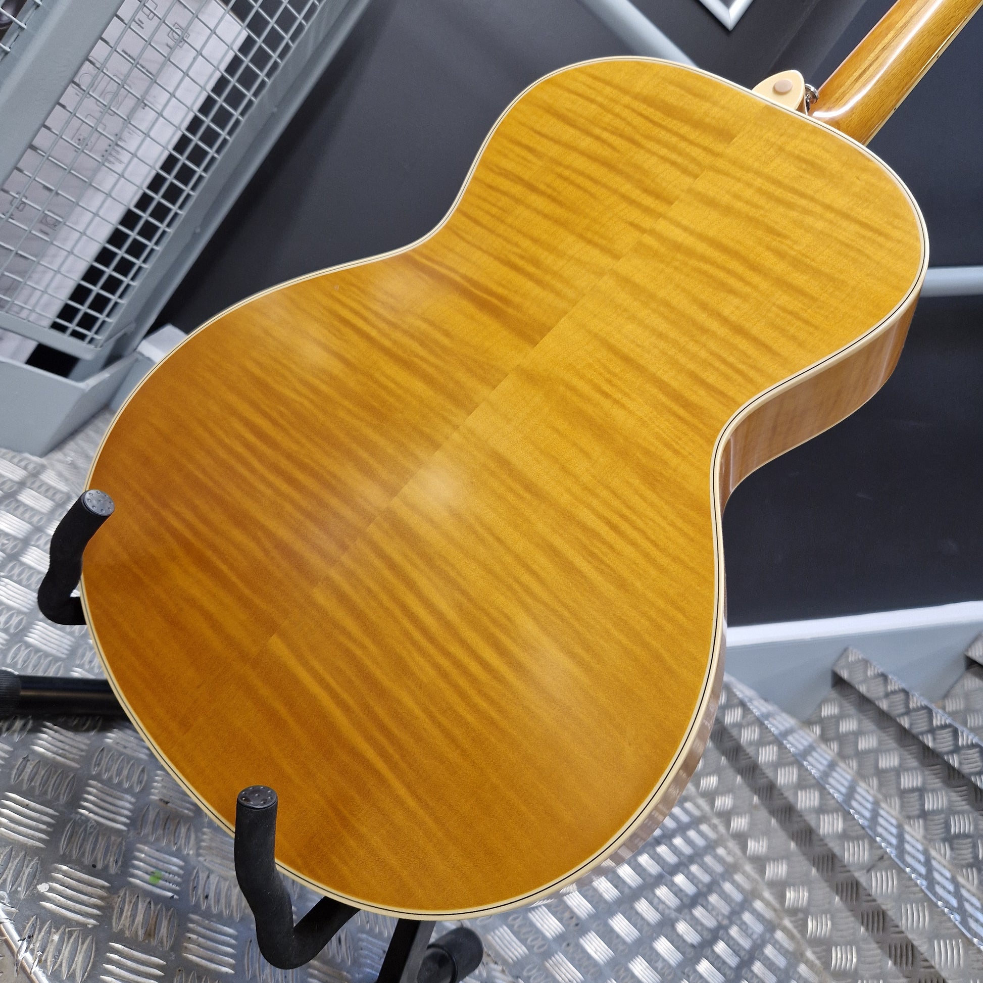 Pre-Owned Epiphone Masterbilt Zenith Classic Electro-Acoustic - Vintage Natural Close-up of the back of a guitar with a glossy, warm honey-toned wood finish, resting on a black stand on a metal diamond plate floor.