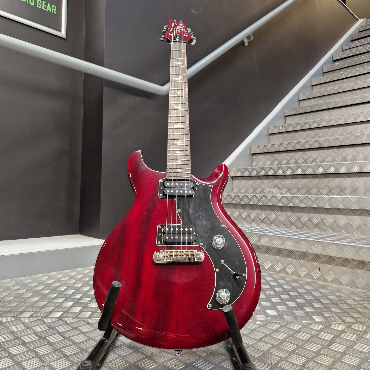 Pre-Owned PRS SE S2 Mira Electric - Wine Red A red electric guitar with a black pickguard and maple neck, displayed on a stand in front of a staircase.