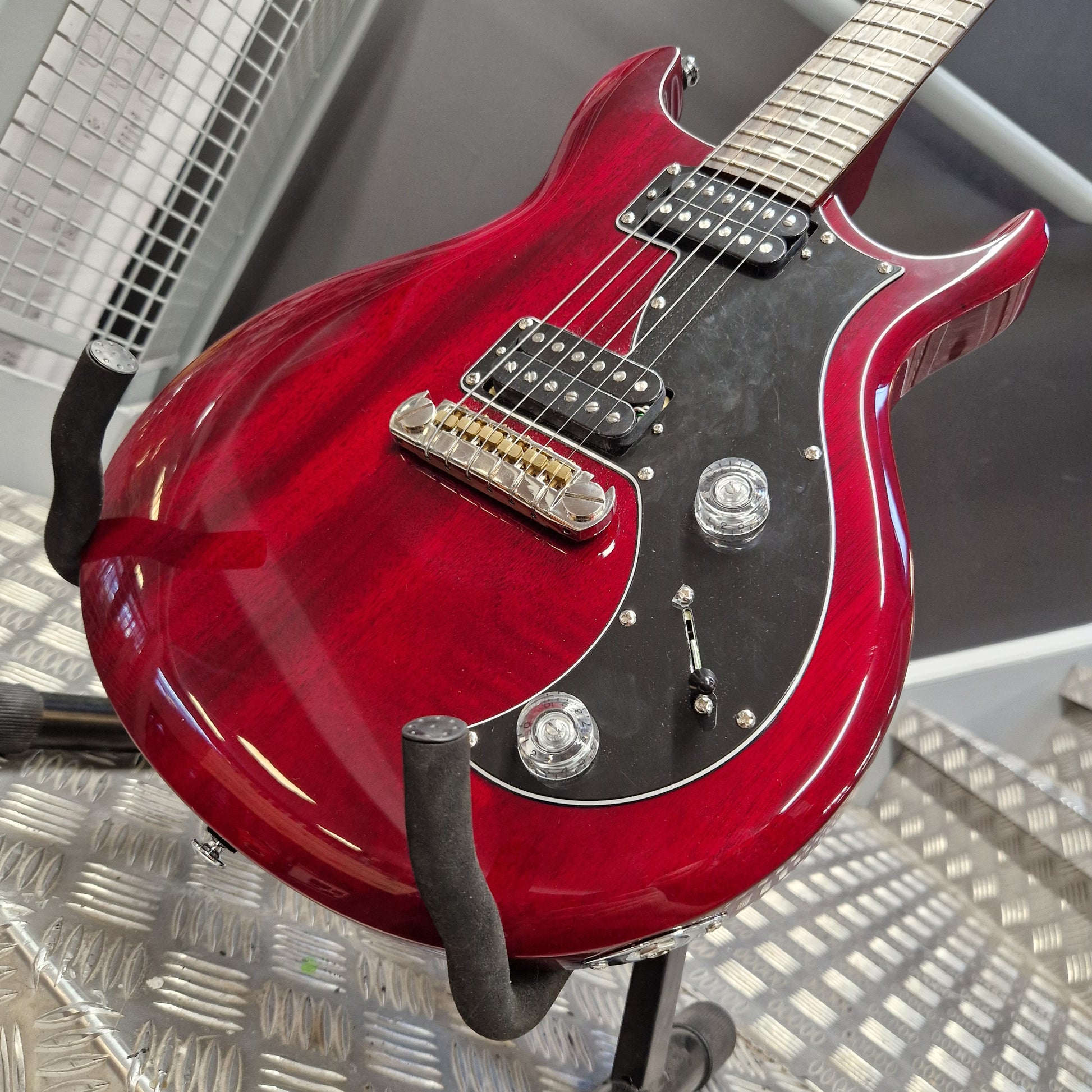 Pre-Owned PRS SE S2 Mira Electric - Wine Red A red electric guitar with a glossy cherry red finish, black pickguard, and chrome hardware, resting on a black guitar stand on a metal diamond plate floor.