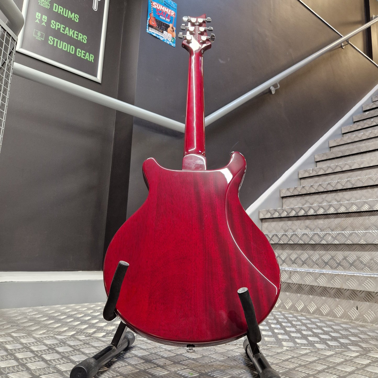 Pre-Owned PRS SE S2 Mira Electric - Wine Red A red electric guitar shown from the back, resting on a black stand in an indoor setting with a staircase and dark gray walls in the background.