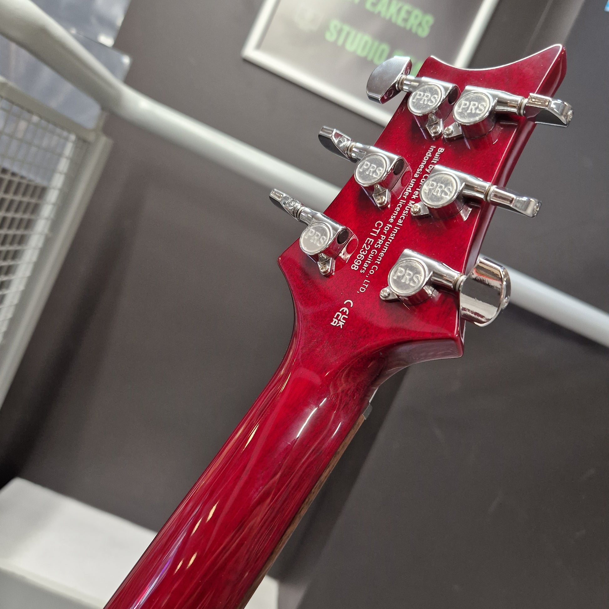 Pre-Owned PRS SE S2 Mira Electric - Wine Red Close-up of the headstock of a glossy red electric guitar, showing six silver tuning pegs and printed model text on the back of the neck, set against a dark gray indoor background.