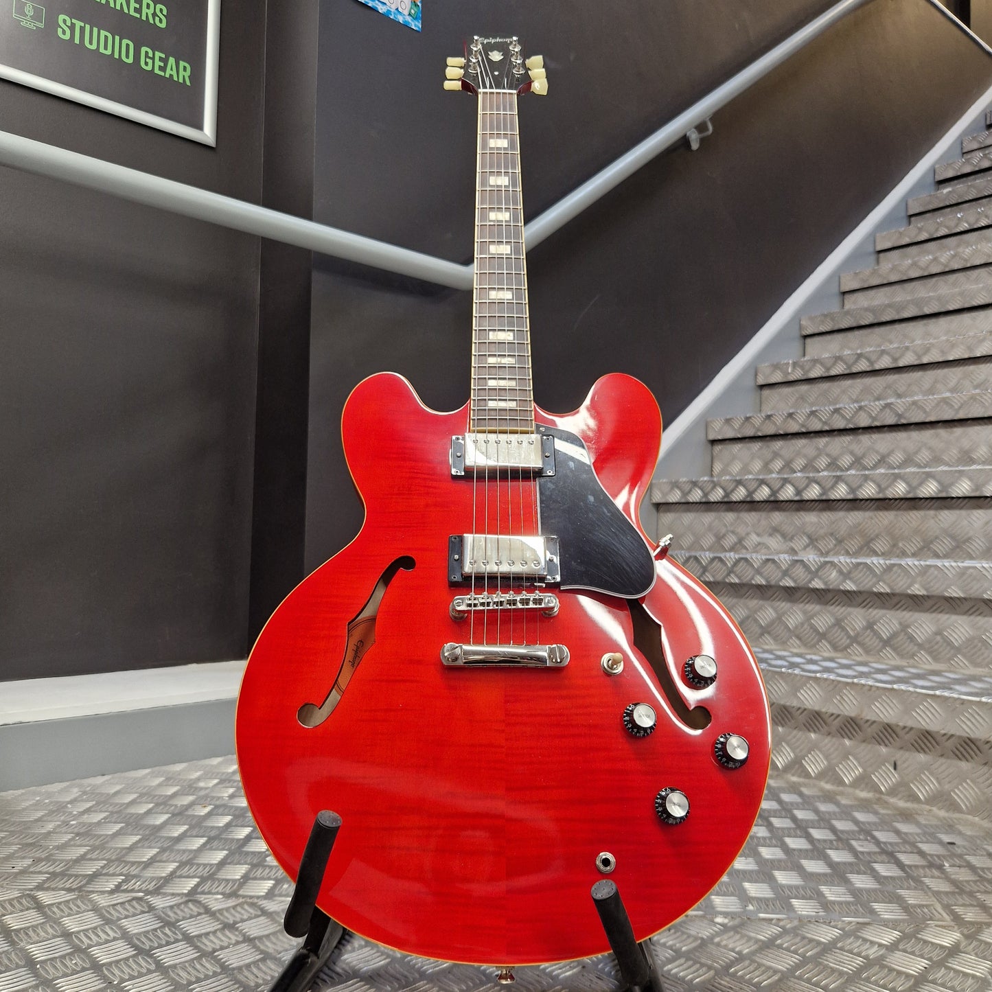 Pre-Owned Epiphone ES-335 Figured Semi-Hollow Electric - Sixties Cherry A red electric guitar with a glossy cherry-red finish, mounted on a stand in front of a staircase and dark wall.