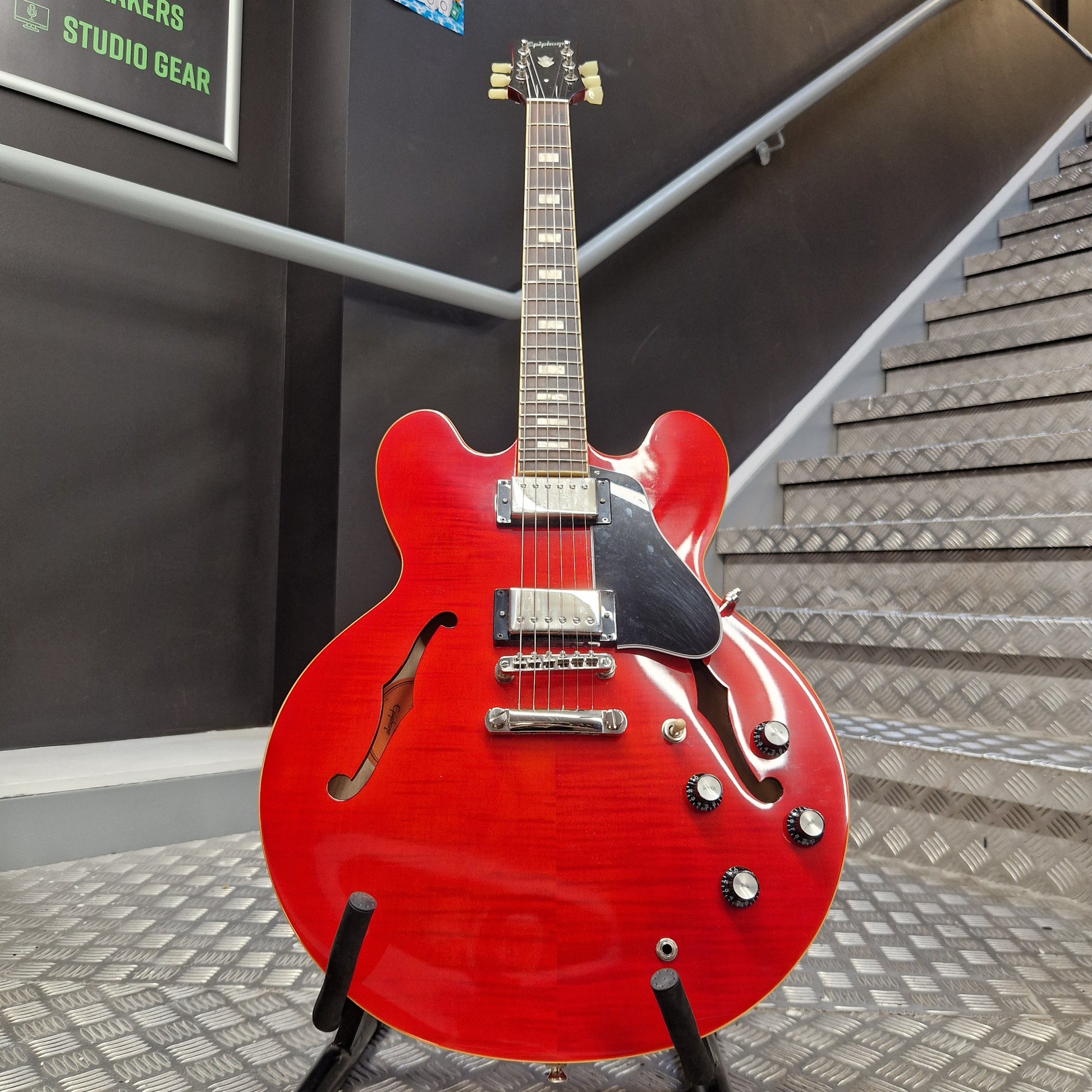 Pre-Owned Epiphone ES-335 Figured Semi-Hollow Electric - Sixties Cherry A red electric guitar with a glossy cherry-red finish, mounted on a stand in front of a staircase and dark wall.