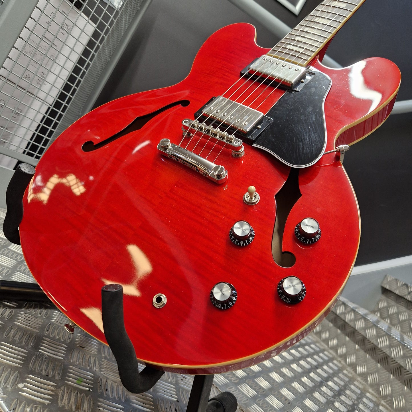 Pre-Owned Epiphone ES-335 Figured Semi-Hollow Electric - Sixties Cherry A red electric guitar with a glossy cherry finish, black pickguard, and chrome hardware, shown resting on a stand in a workshop setting.