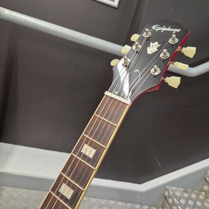 Pre-Owned Epiphone ES-335 Figured Semi-Hollow Electric - Sixties Cherry Close-up of an Epiphone acoustic guitar headstock and neck, showing the brand name, six tuning pegs, and dark wood fretboard against a dark gray wall.