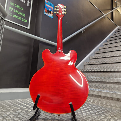 Pre-Owned Epiphone ES-335 Figured Semi-Hollow Electric - Sixties Cherry A red electric guitar shown from the back, standing on a black display stand in an indoor setting with a staircase and dark wall in the background.