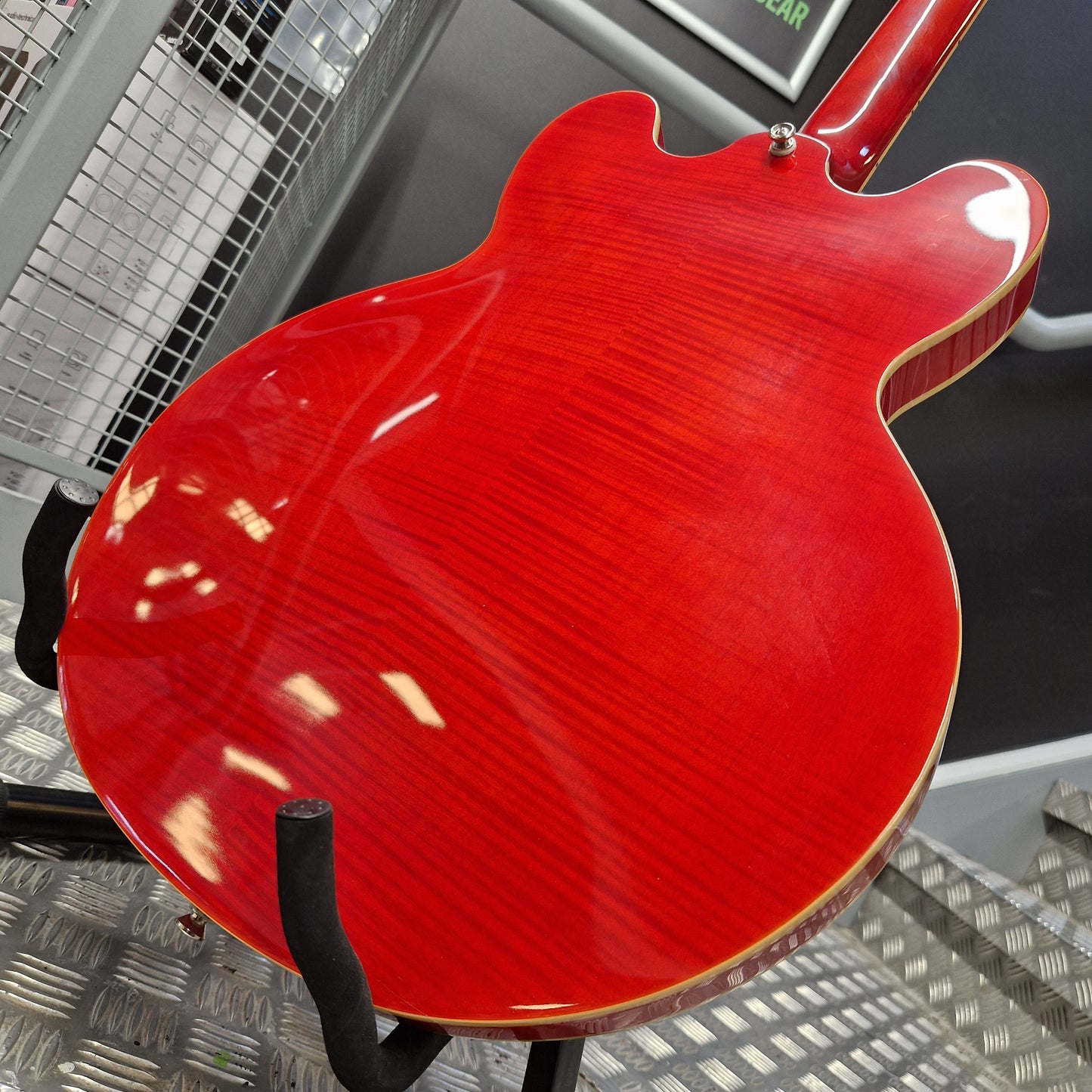 Pre-Owned Epiphone ES-335 Figured Semi-Hollow Electric - Sixties Cherry Close-up of the glossy red back of an electric guitar on a black stand, with a metal workshop floor and mesh background visible.
