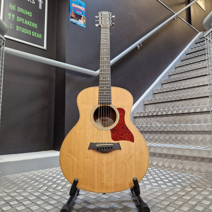 Pre-Owned Taylor GS Mini-e Electro-Acoustic - Limited Edition Quilted Sapele A natural wood acoustic guitar with a red glittery pickguard stands on a black stand in front of a staircase and dark gray wall.