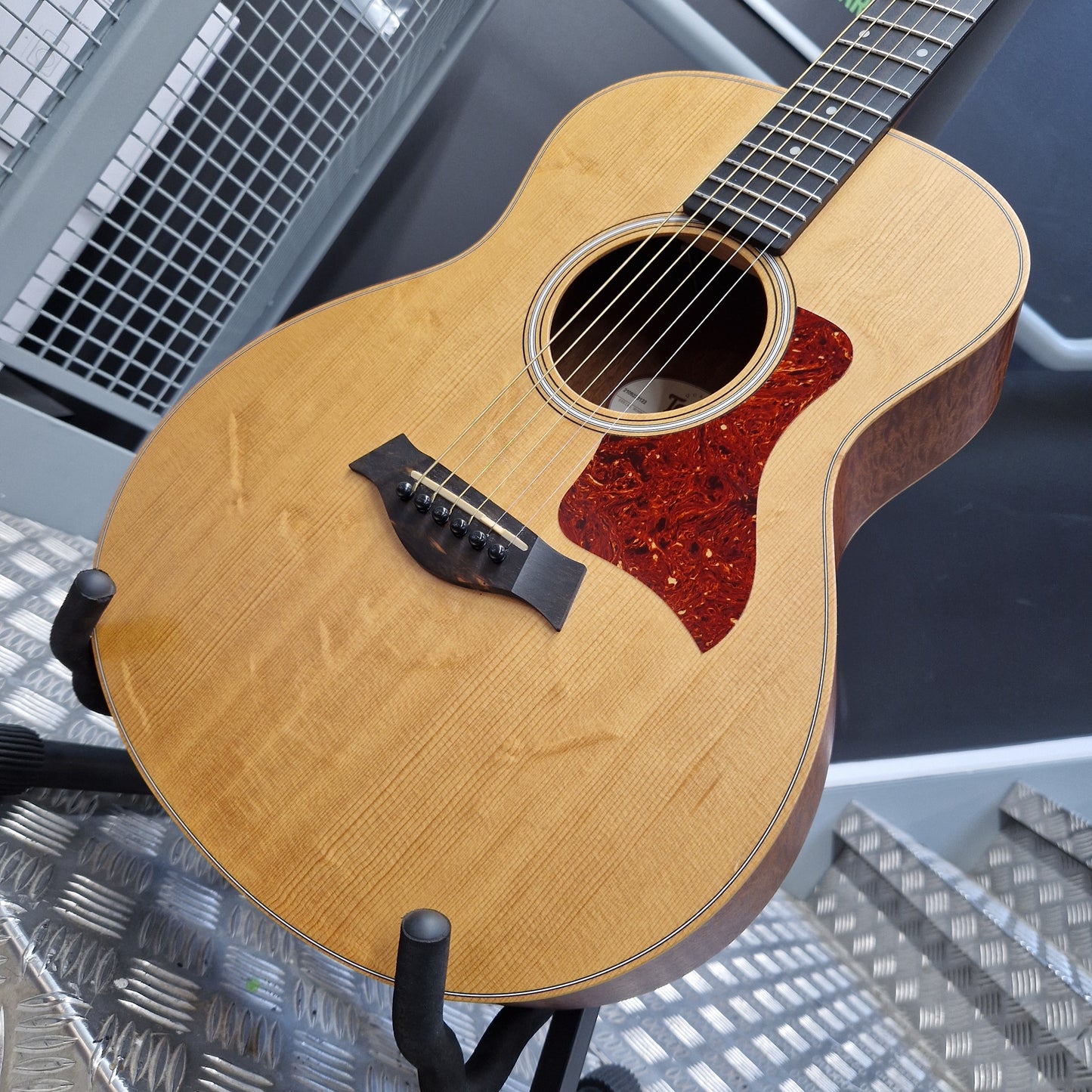Pre-Owned Taylor GS Mini-e Electro-Acoustic - Limited Edition Quilted Sapele An acoustic guitar with a natural wood body and a red glittery pickguard, shown resting on a stand in what appears to be a workshop or studio setting.