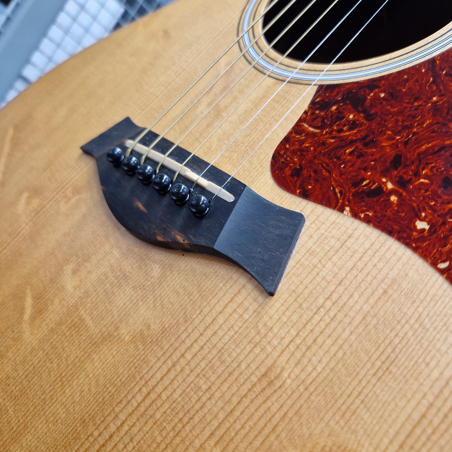 Pre-Owned Taylor GS Mini-e Electro-Acoustic - Limited Edition Quilted Sapele Close-up of an acoustic guitar body showing the natural wood grain, black bridge, strings, and a reddish-brown tortoiseshell-style pickguard.