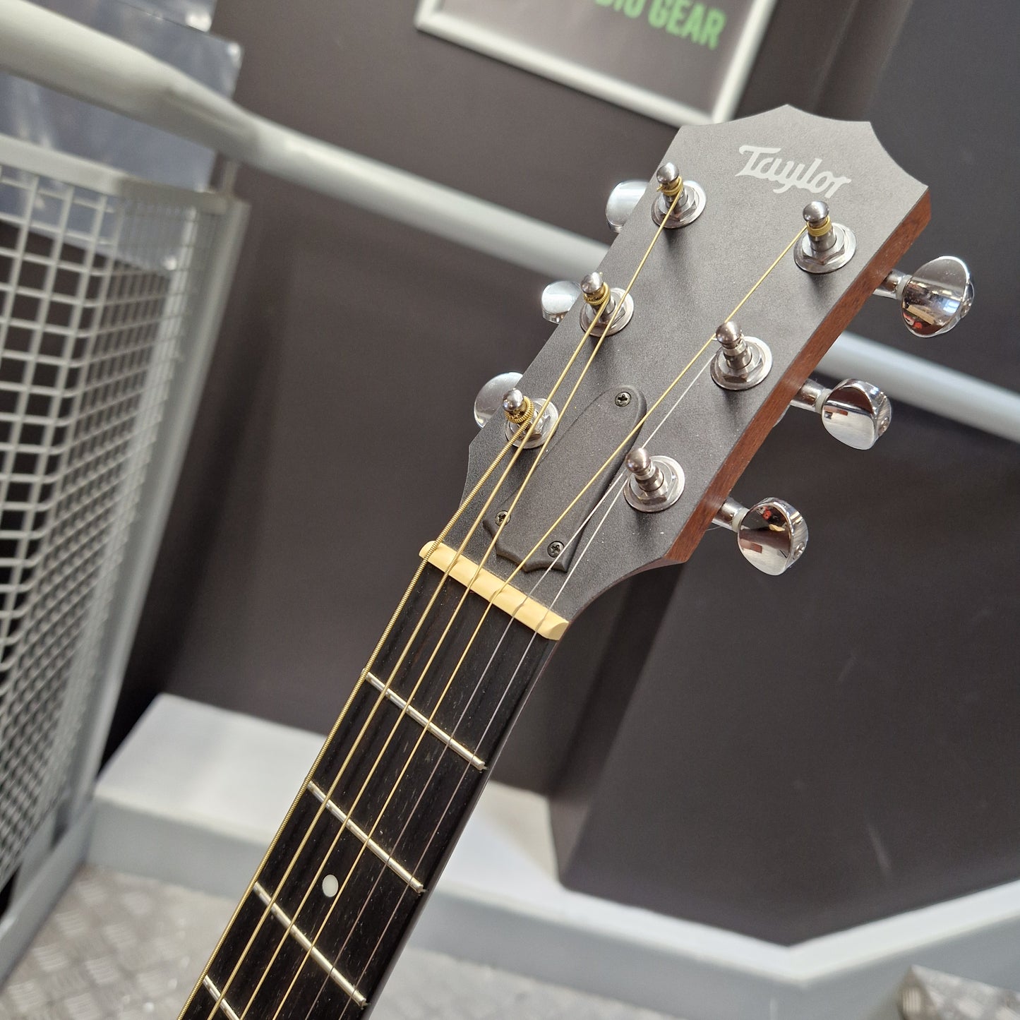 Pre-Owned Taylor GS Mini-e Electro-Acoustic - Limited Edition Quilted Sapele Close-up of a Taylor acoustic guitar headstock and upper neck, showing six silver tuning pegs, gold strings, and a dark gray headstock against an indoor background.