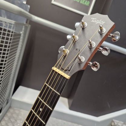 Pre-Owned Taylor GS Mini-e Electro-Acoustic - Limited Edition Quilted Sapele Close-up of a Taylor acoustic guitar headstock and upper neck, showing six silver tuning pegs, gold strings, and a dark gray headstock against an indoor background.