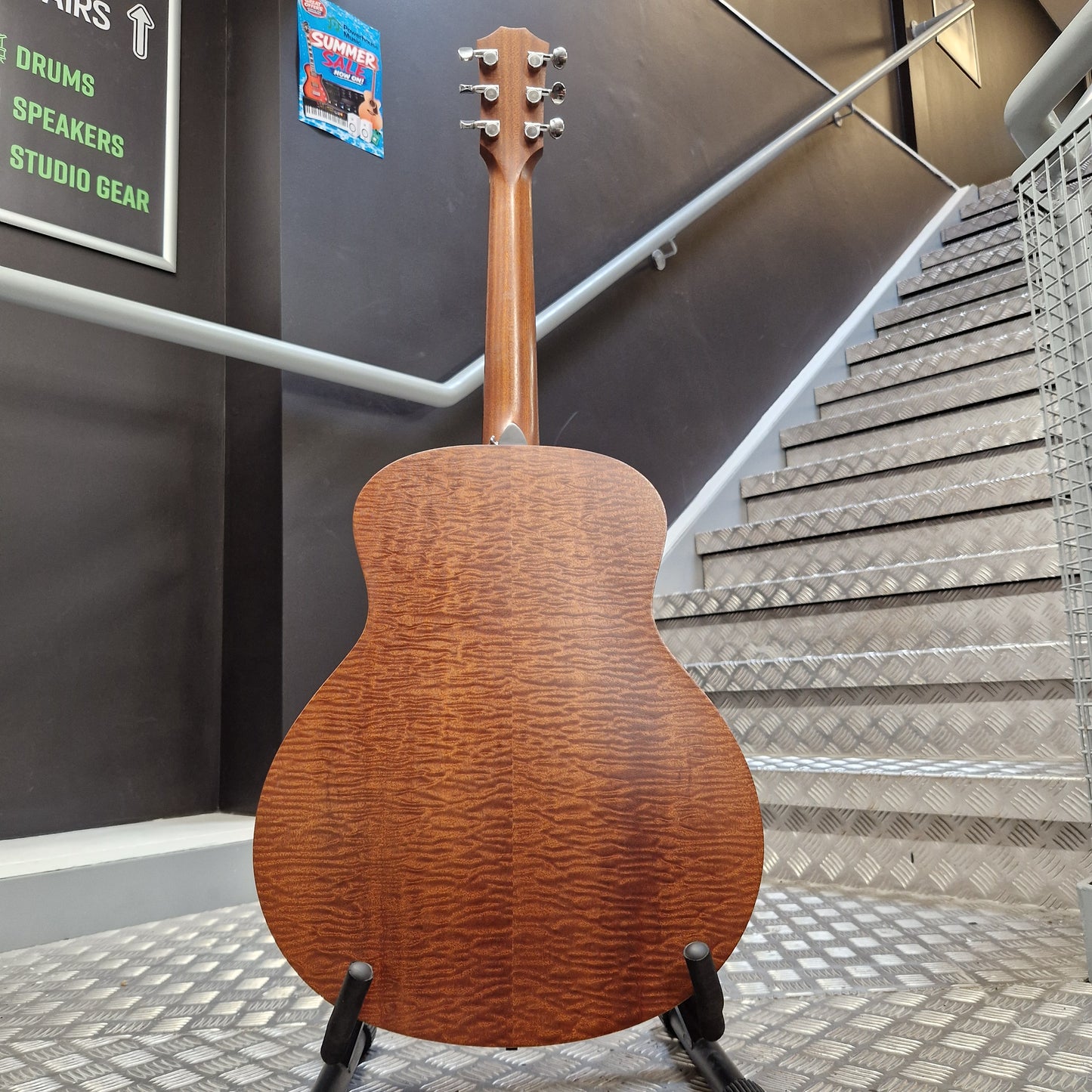 Pre-Owned Taylor GS Mini-e Electro-Acoustic - Limited Edition Quilted Sapele The back of a wooden acoustic guitar with a glossy flamed maple top, displayed on a stand in front of a staircase.