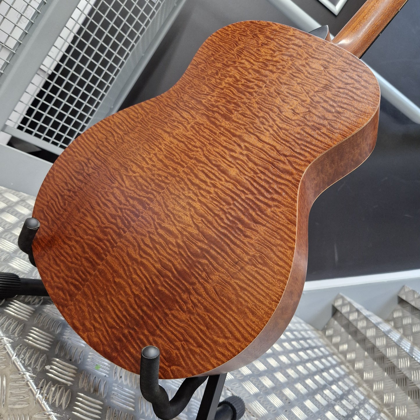 Pre-Owned Taylor GS Mini-e Electro-Acoustic - Limited Edition Quilted Sapele Close-up of the back of an acoustic guitar with a rich, glossy reddish-brown wood finish and prominent grain pattern, resting on a black stand.