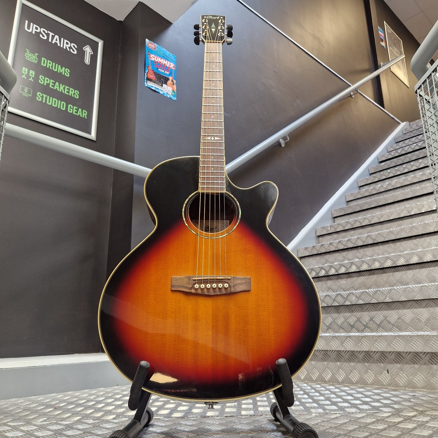 Pre-Owned Cort SFX6R Electro-Acoustic - 3-Tone Sunburst A cutaway acoustic-electric guitar with a sunburst finish, displayed on a stand in front of a staircase.