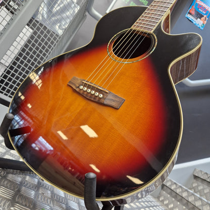 Pre-Owned Cort SFX6R Electro-Acoustic - 3-Tone Sunburst An acoustic-electric guitar with a sunburst finish, resting on a stand in what appears to be a showroom or workshop.