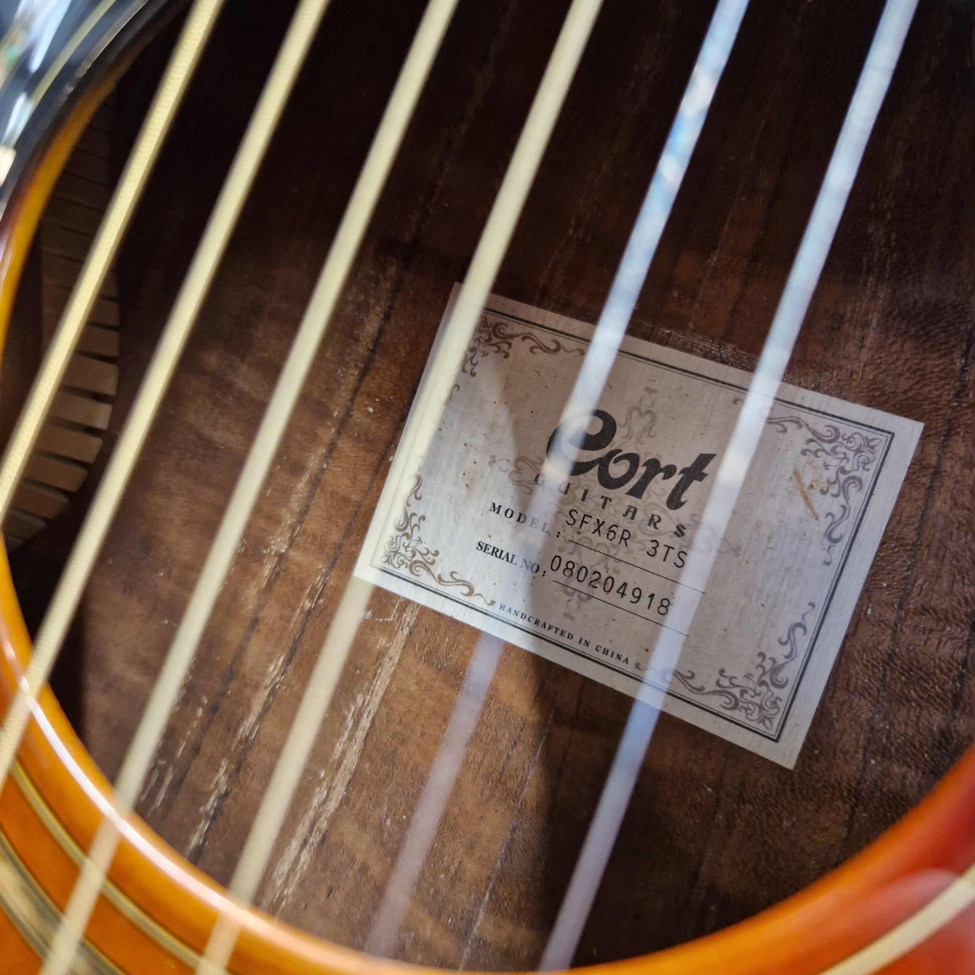 Pre-Owned Cort SFX6R Electro-Acoustic - 3-Tone Sunburst Close-up of the inside of an acoustic guitar body showing the wood grain, sound hole, strings, and a visible Cort guitar label.