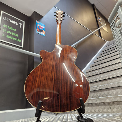 Pre-Owned Cort SFX6R Electro-Acoustic - 3-Tone Sunburst The back of a glossy brown acoustic guitar on a stand, positioned in front of a staircase and dark gray walls.