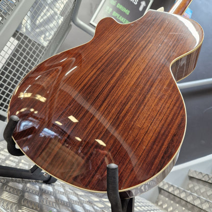 Pre-Owned Cort SFX6R Electro-Acoustic - 3-Tone Sunburst Close-up of the glossy back of an electric guitar showing a rich dark brown wood grain finish, displayed on a stand.