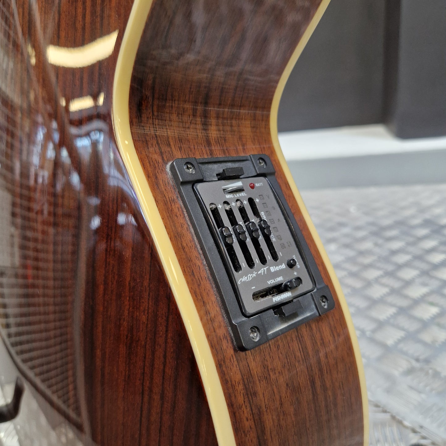Pre-Owned Cort SFX6R Electro-Acoustic - 3-Tone Sunburst Close-up of the lower bout corner of an acoustic guitar with a glossy dark brown wood finish, cream binding, and a black magnetic volume control plate.