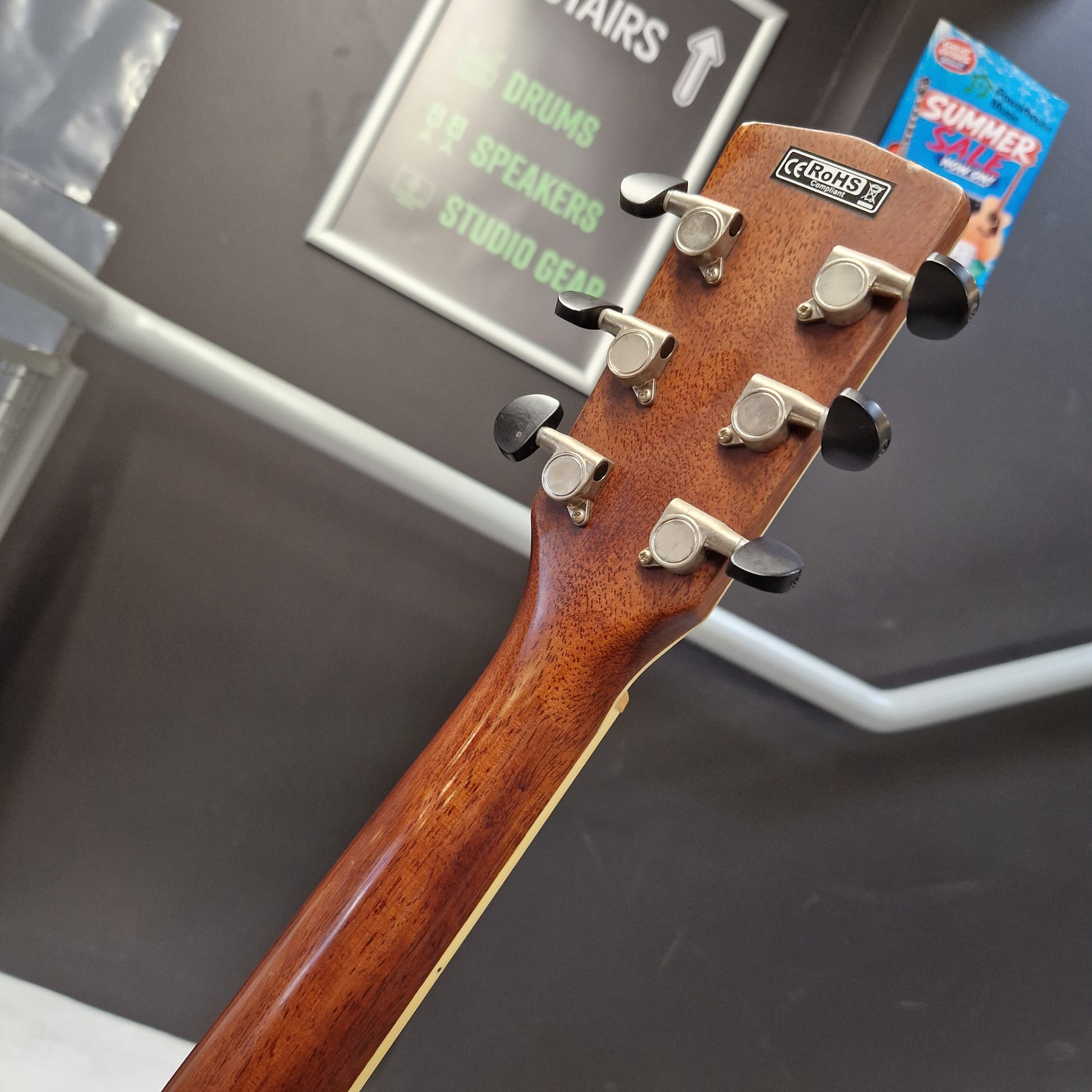 Pre-Owned Cort SFX6R Electro-Acoustic - 3-Tone Sunburst Close-up of the headstock of a Crets F acoustic guitar, showing the glossy brown wood grain and six silver tuning pegs with black caps, set against a dark gray background with store signage.
