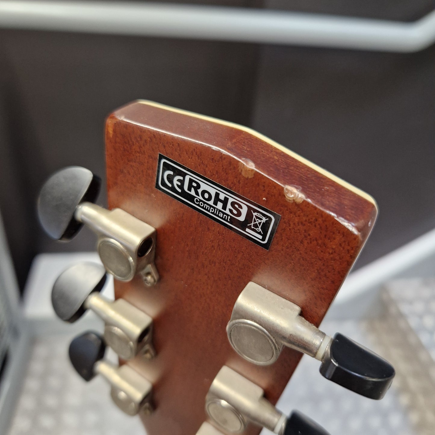Pre-Owned Cort SFX6R Electro-Acoustic - 3-Tone Sunburst Close-up of the headstock of a guitar, showing a reddish-brown wood finish, silver tuning pegs, and a black and white CE RoHS compliant sticker.
