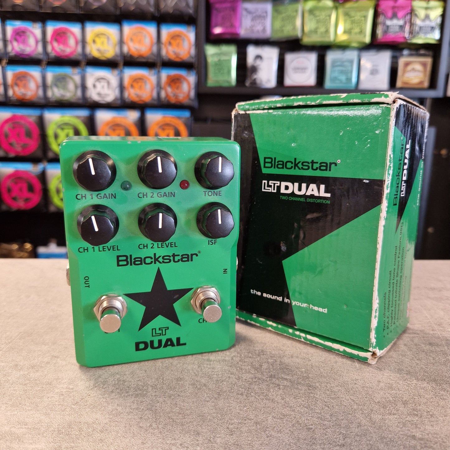 Pre-Owned Blackstar LT-DUAL Two-Channel Distortion Pedal A Blackstar LT Dual guitar effects pedal in green with black knobs and silver footswitches, shown next to its original green and black retail box on a tabletop. The background shows a store shelf filled with colorful items.