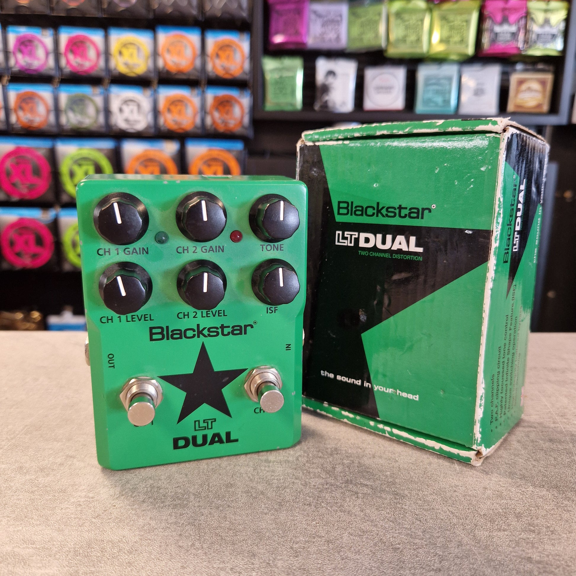 Pre-Owned Blackstar LT-DUAL Two-Channel Distortion Pedal A Blackstar LT Dual guitar effects pedal in green with black knobs and silver footswitches, shown next to its original green and black retail box on a tabletop. The background shows a store shelf filled with colorful items.