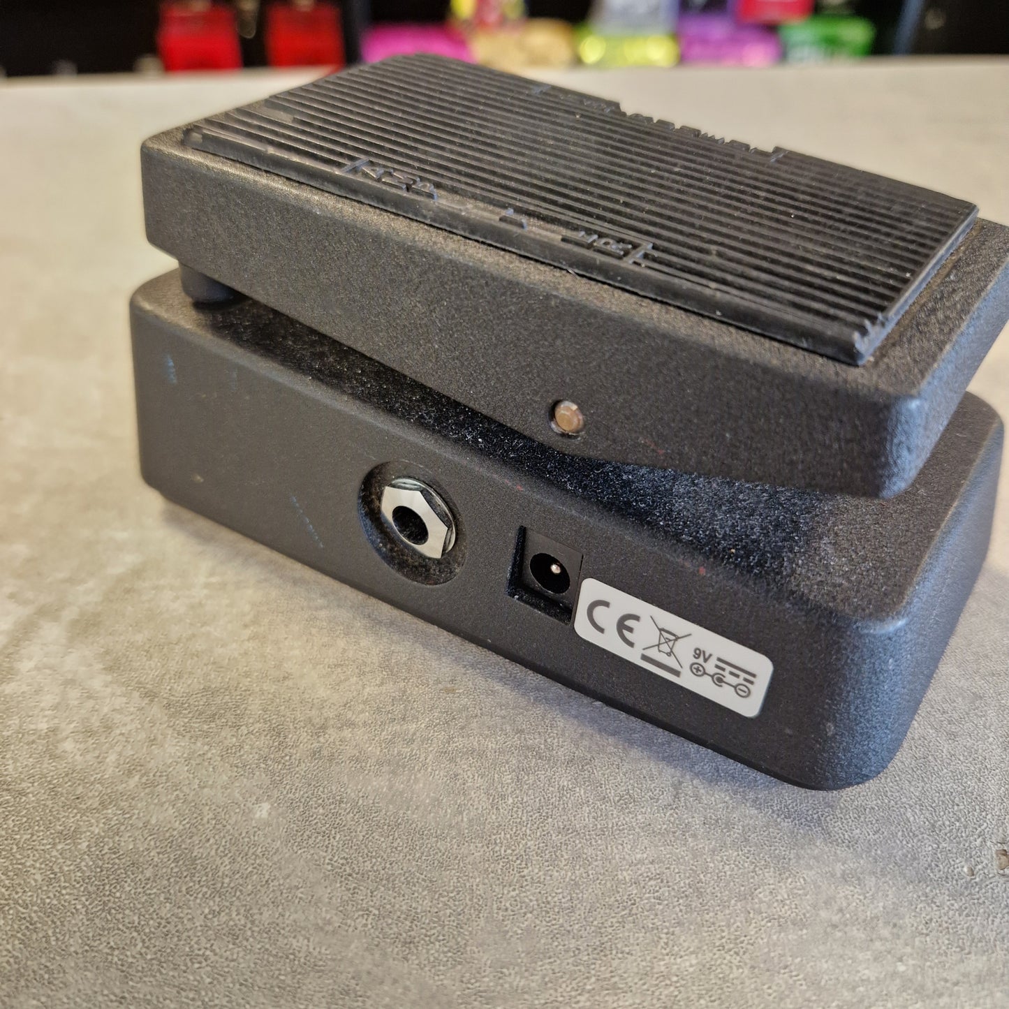 Pre-Owned Dunlop CBM95 Cry Baby Mini Wah Pedal A black guitar effects pedal with a ribbed metal footplate, photographed from the side to show the jack input and power output jacks on a gray textured surface.