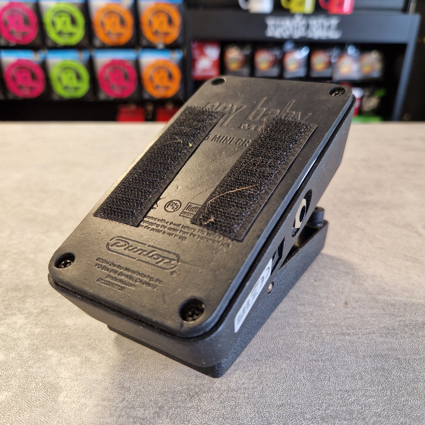 Pre-Owned Dunlop CBM95 Cry Baby Mini Wah Pedal A black Dunlop baby cradle base with velcro pads and mounting hardware, photographed on a gray tabletop with a blurred store background.