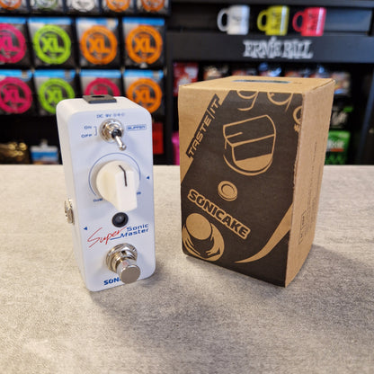 Pre-Owned Sonicake Super Sonic Master Boost Pedal A white Sonicake Super Sonic Master guitar effects pedal with silver controls stands next to its brown cardboard box on a gray tabletop. The background shows a blurred music store shelf with colorful knobs and mugs.
