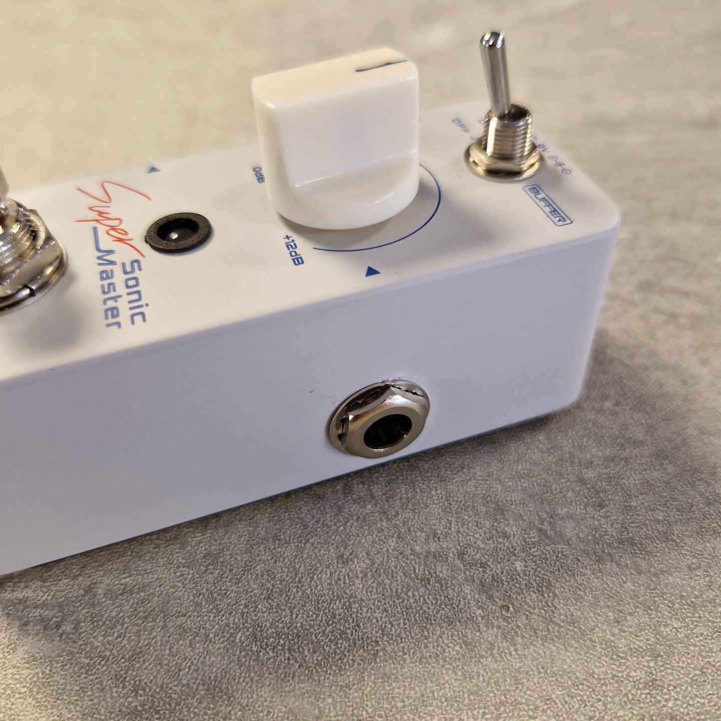 Pre-Owned Sonicake Super Sonic Master Boost Pedal A close-up of a white Sonic Master Super Master guitar effects pedal, showing the white volume knob, silver toggle switch, and metal jack connectors on a textured gray surface.