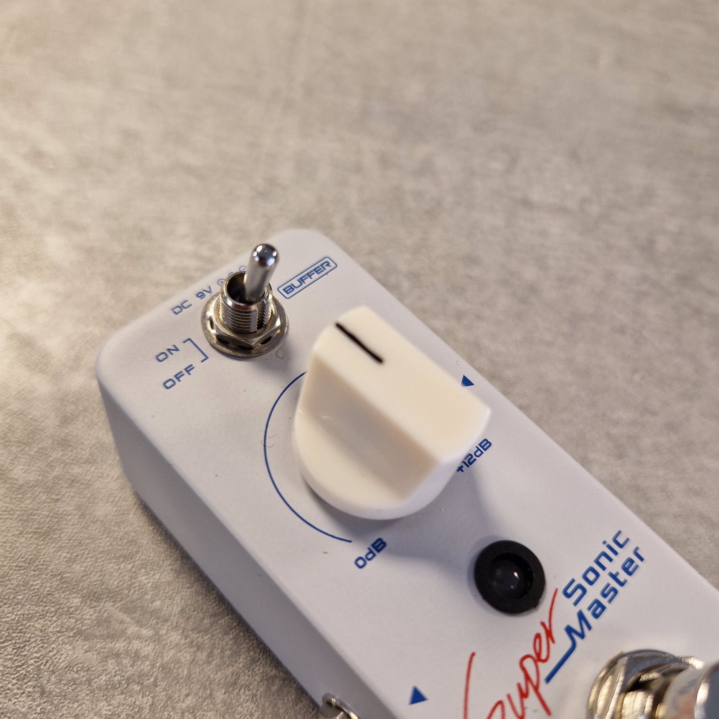 Pre-Owned Sonicake Super Sonic Master Boost Pedal A close-up of a white Super Sonic Master guitar effects pedal with a white rotary volume knob, silver switch, and blue/red printed markings on a textured gray surface.