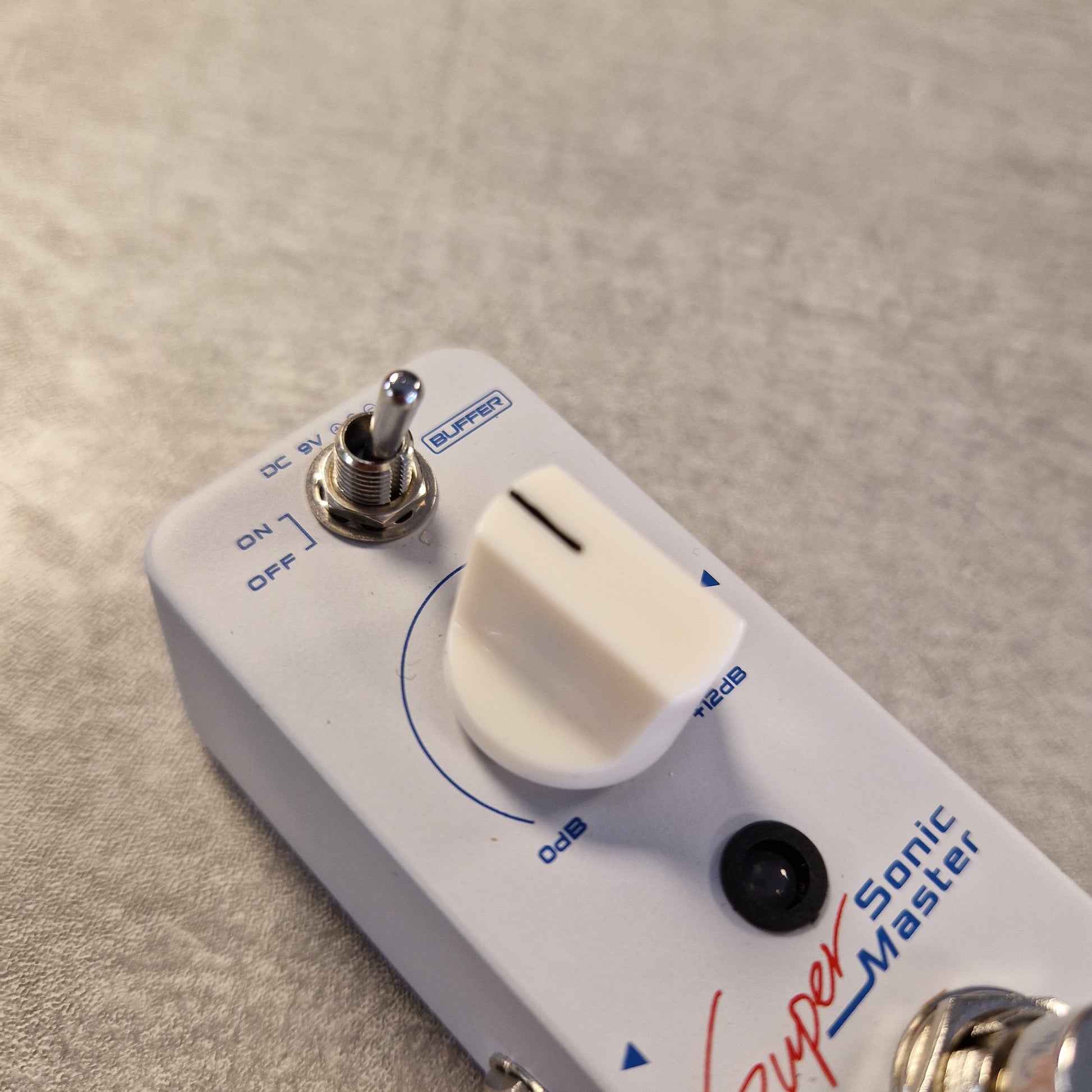 Pre-Owned Sonicake Super Sonic Master Boost Pedal A close-up of a white Super Sonic Master guitar effects pedal with a white rotary volume knob, silver switch, and blue/red printed markings on a textured gray surface.