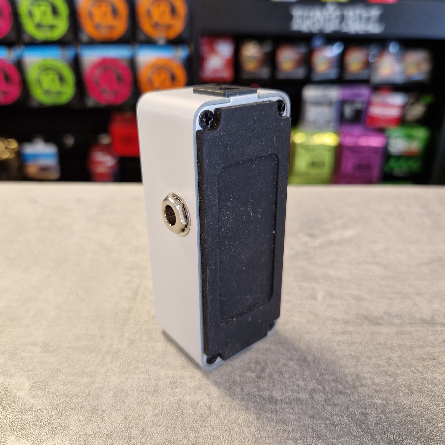 Pre-Owned Sonicake Super Sonic Master Boost Pedal A small rectangular electronic device with a white outer shell and a black textured back panel, standing on a gray tabletop with a blurred retail display in the background.