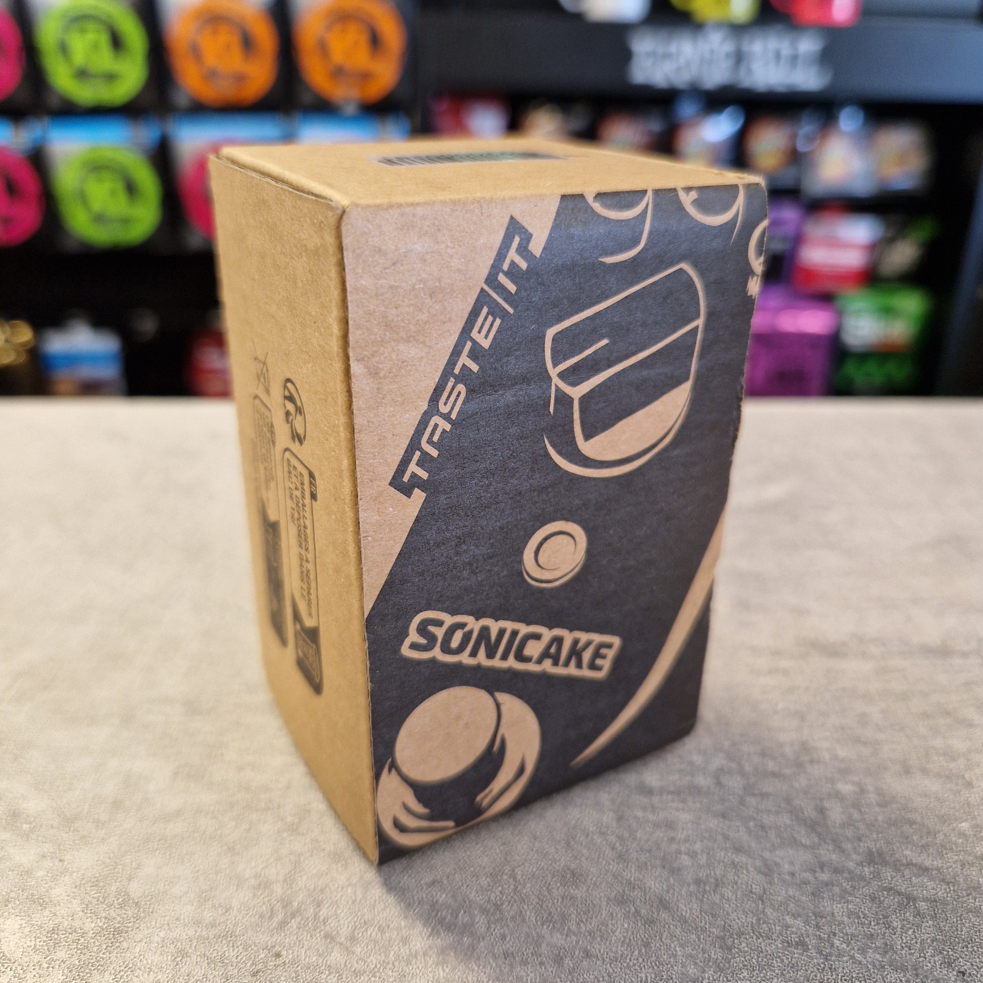 Pre-Owned Sonicake Super Sonic Master Boost Pedal A cardboard product box for Sonicake, featuring a dark gray printed design with coffee cup illustrations and the brand name 'SONICAKE' and slogan 'TASTE IT', set on a gray tabletop with a blurred store background.