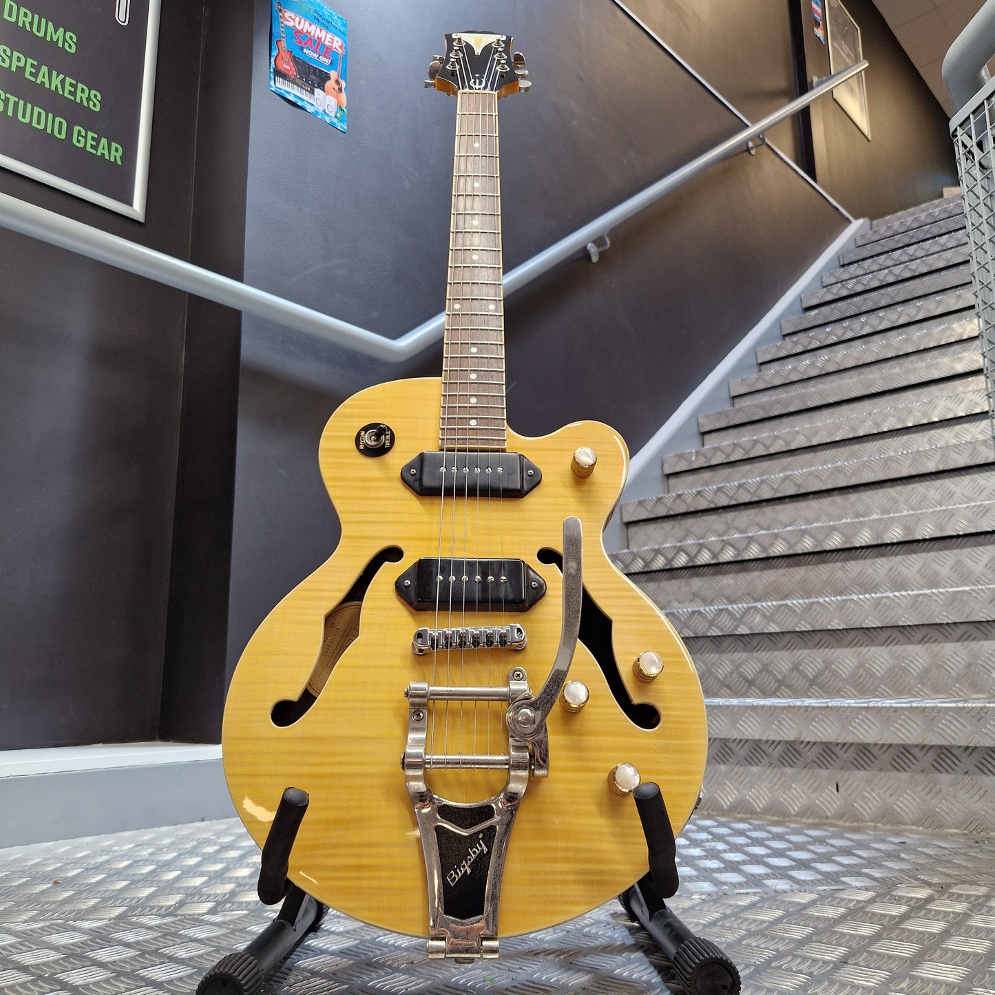 Pre-Owned Epiphone Wildkat Hollow-Body Electric - Natural Amber A yellow-finish electric guitar with a silver tremolo bridge and black pickups, displayed on a stand in front of a staircase.