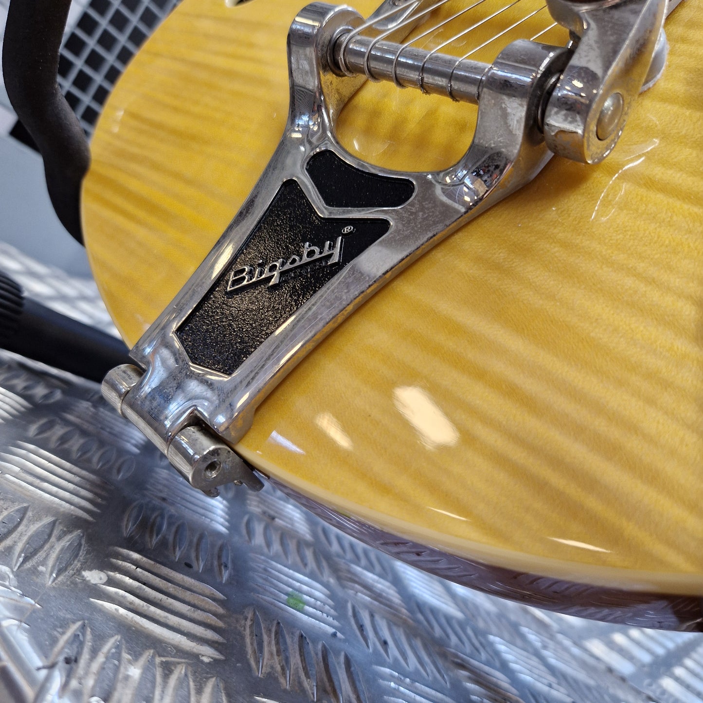 Pre-Owned Epiphone Wildkat Hollow-Body Electric - Natural Amber Close-up of a shiny silver Bigby brand guitar tailpiece mounted on a yellow guitar body over a textured silver metal surface.