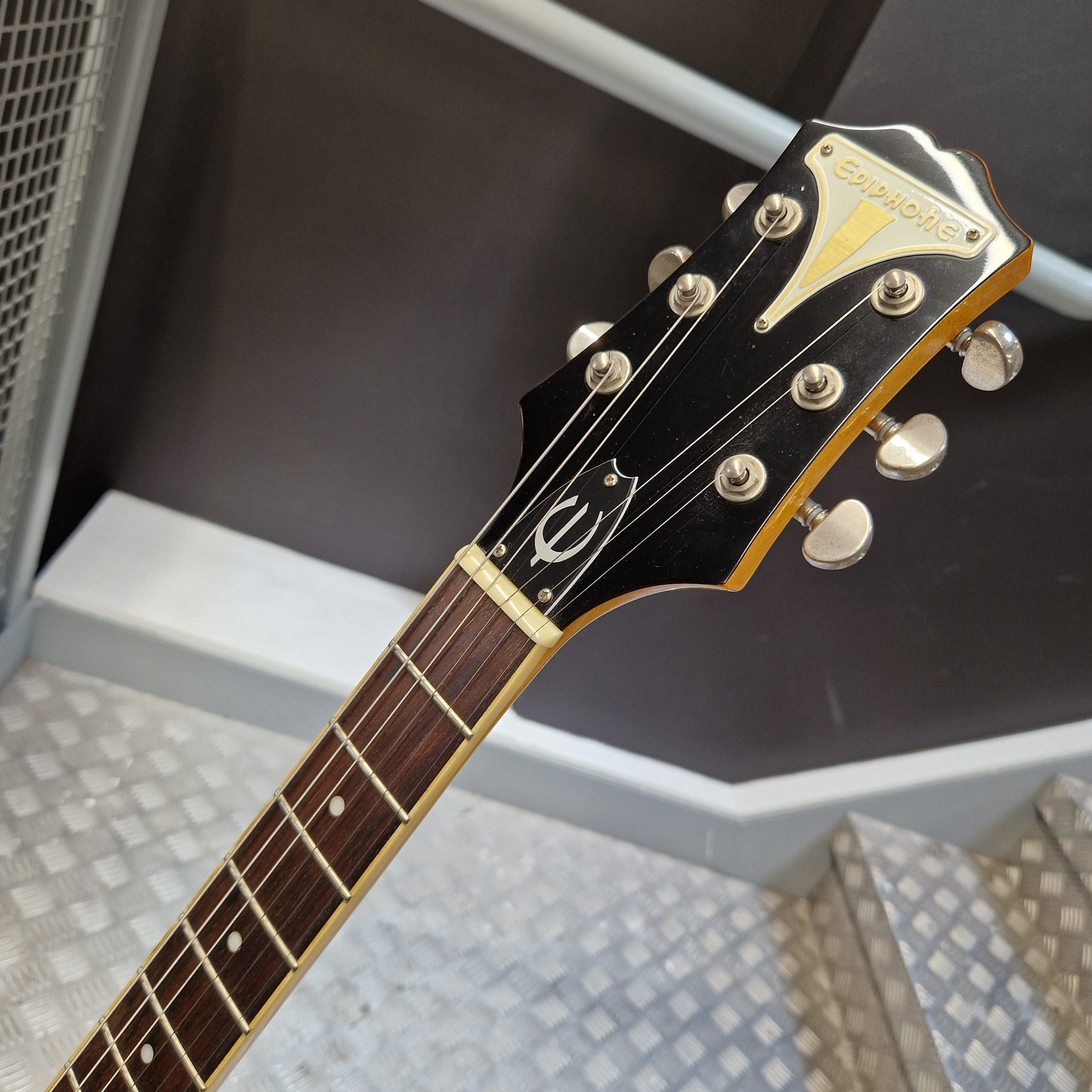 Pre-Owned Epiphone Wildkat Hollow-Body Electric - Natural Amber Close-up of a Gibson electric guitar headstock and neck, showing the black headstock with gold hardware and a dark fretboard, photographed indoors against a gray wall and metal steps.