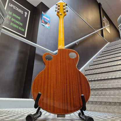 Pre-Owned Epiphone Wildkat Hollow-Body Electric - Natural Amber A Gibson Les Paul Custom electric guitar shown from the back, featuring a glossy orange-brown wood finish and a bright yellow headstock, displayed on a stand in a stairwell.