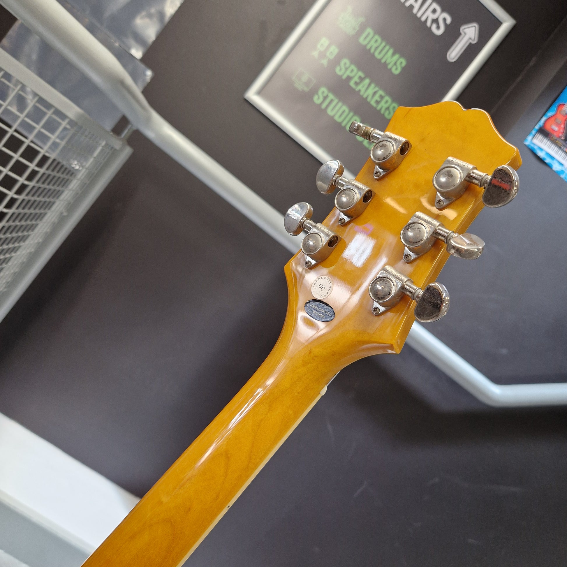 Pre-Owned Epiphone Wildkat Hollow-Body Electric - Natural Amber Close-up of the headstock of a yellow-orange acoustic guitar, showing six silver tuning pegs and a glossy maple neck, photographed in a store or studio setting.