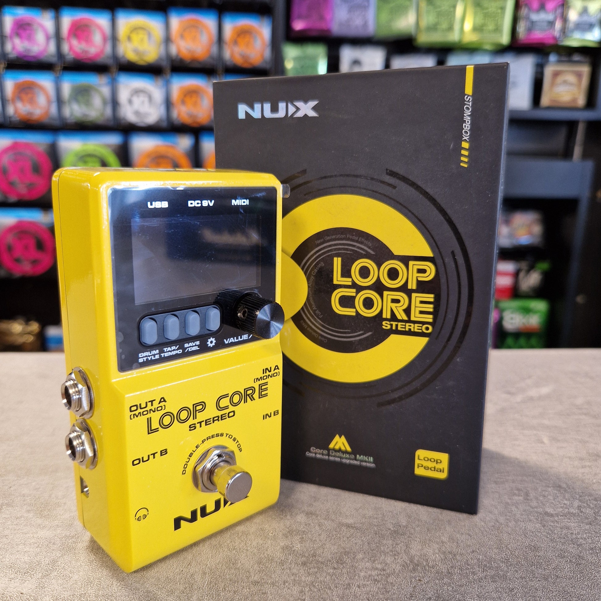 Pre-Owned Nux Loop Core Stereo Looper Pedal A yellow NUIX Loop Core Stereo guitar pedal in the foreground with its black retail box behind it, photographed on a tabletop with a blurred store background.