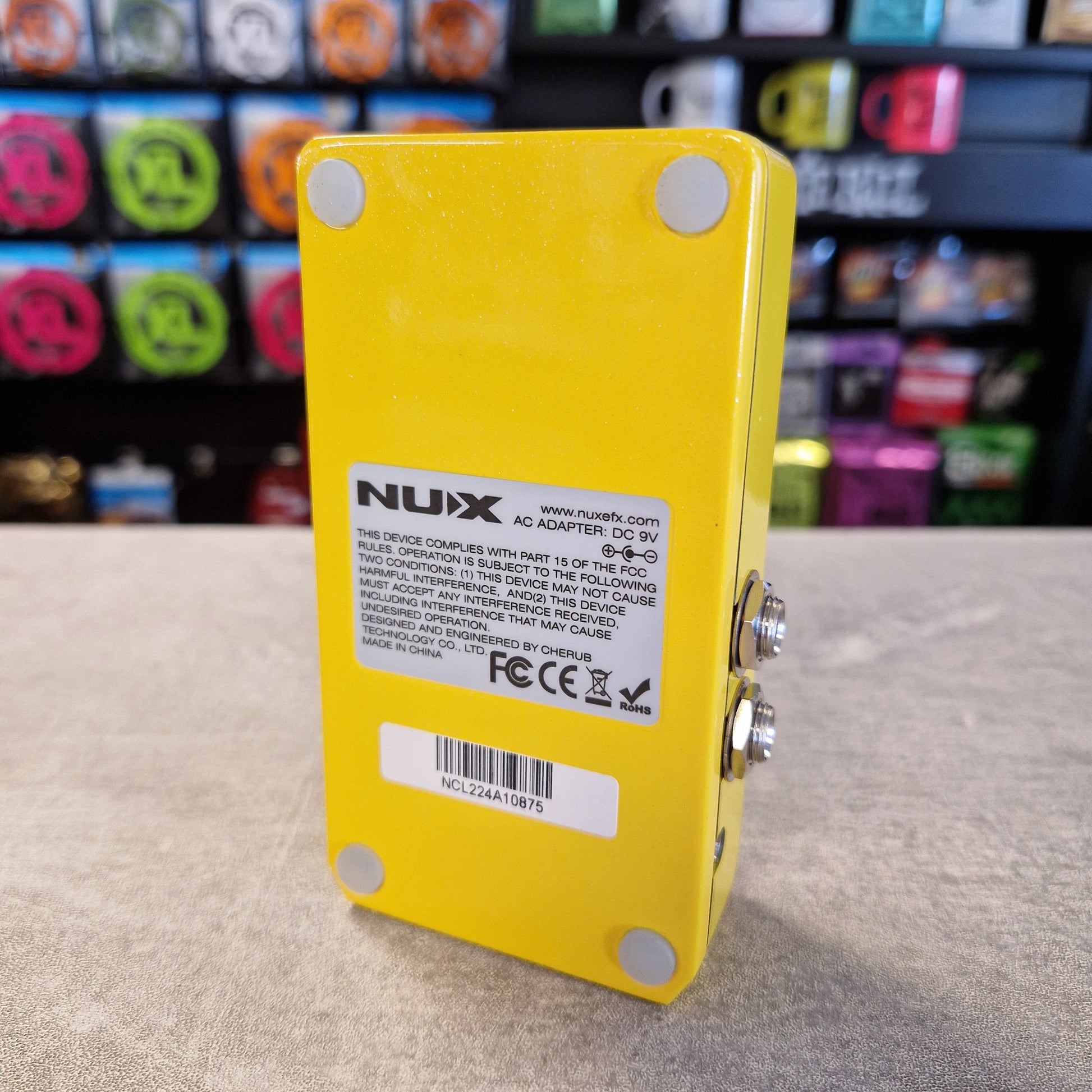Pre-Owned Nux Loop Core Stereo Looper Pedal A bright yellow NUX guitar effects adapter pedal shown from the back, featuring a white specification label and silver metal input/output jacks, set against a blurred background of colorful guitar headstocks.