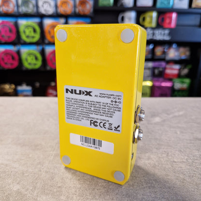 Pre-Owned Nux Loop Core Stereo Looper Pedal A bright yellow NUX guitar effects adapter pedal shown from the back, featuring a white specification label and silver metal input/output jacks, set against a blurred background of colorful guitar headstocks.