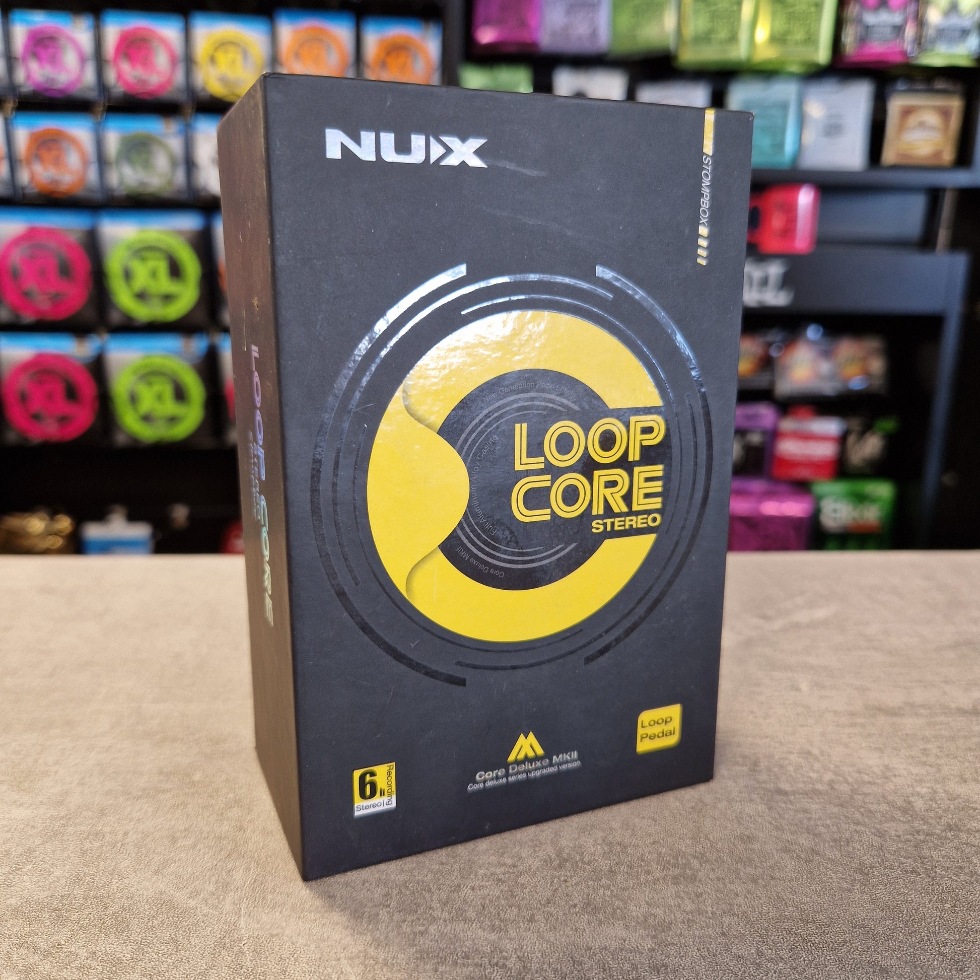 Pre-Owned Nux Loop Core Stereo Looper Pedal A black software box for NUX Loop Core Stereo, featuring a bold yellow circular logo, standing on a beige surface with a blurred background of colorful items.